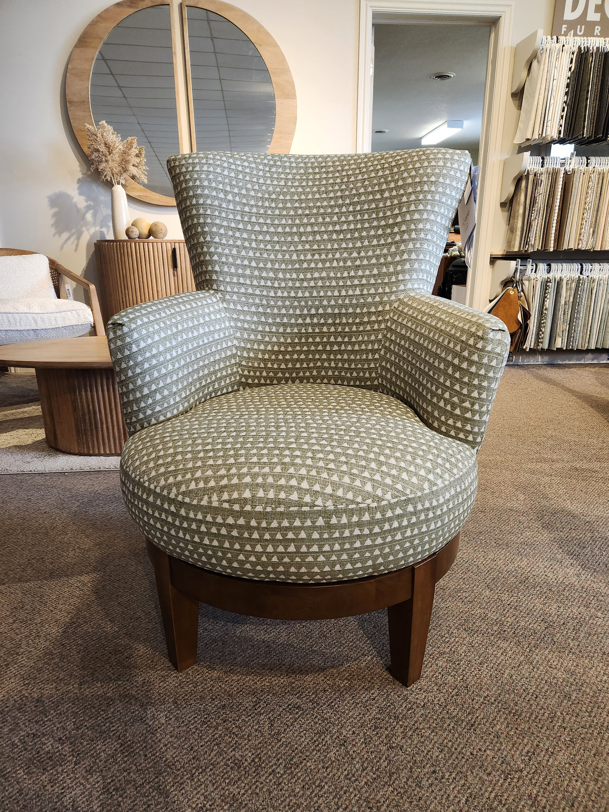 The 2968E Justine Swivel Chair by Best, features green patterned fabric with wooden legs, in a showroom with samples and a mirror.