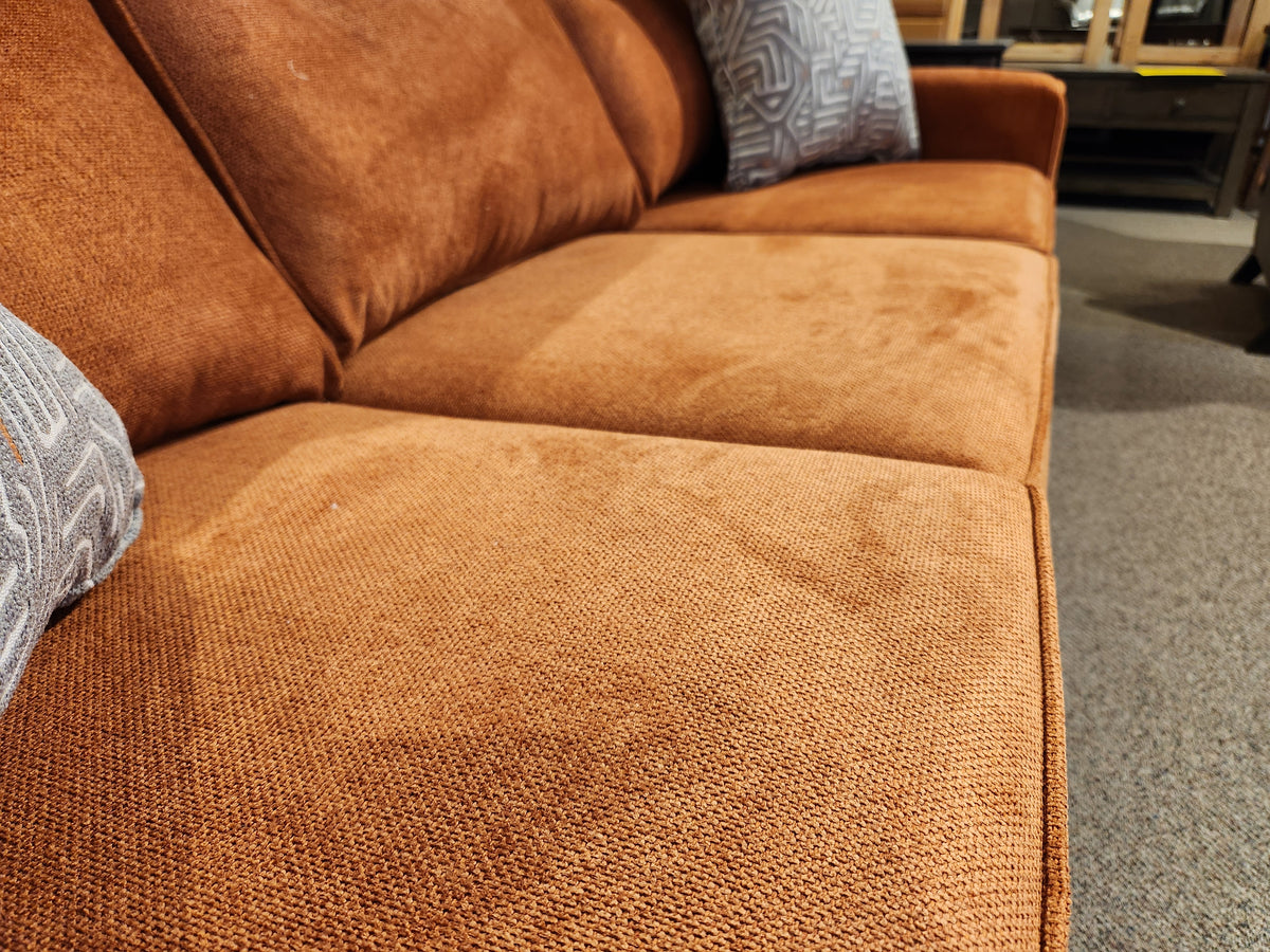 Close-up of the Trendline 4731 Sofa, a Canadian-made piece featuring orange fabric with a button-tufted back cushion, highlighted by two patterned cushions, and set on a carpeted floor.