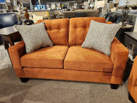 The Trendline 4731 Loveseat, featuring an orange design with button-tufted detailing and two patterned pillows, is on display in a furniture store setting, highlighting a wide selection of fabrics.