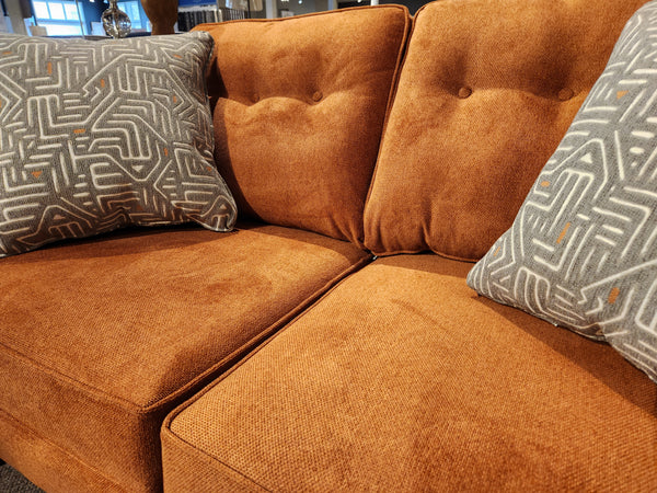 The orange-brown 4731 Loveseat by Trendline features button-tufted details and comes with geometric-patterned gray pillows, beautifully displayed in a showroom setting.