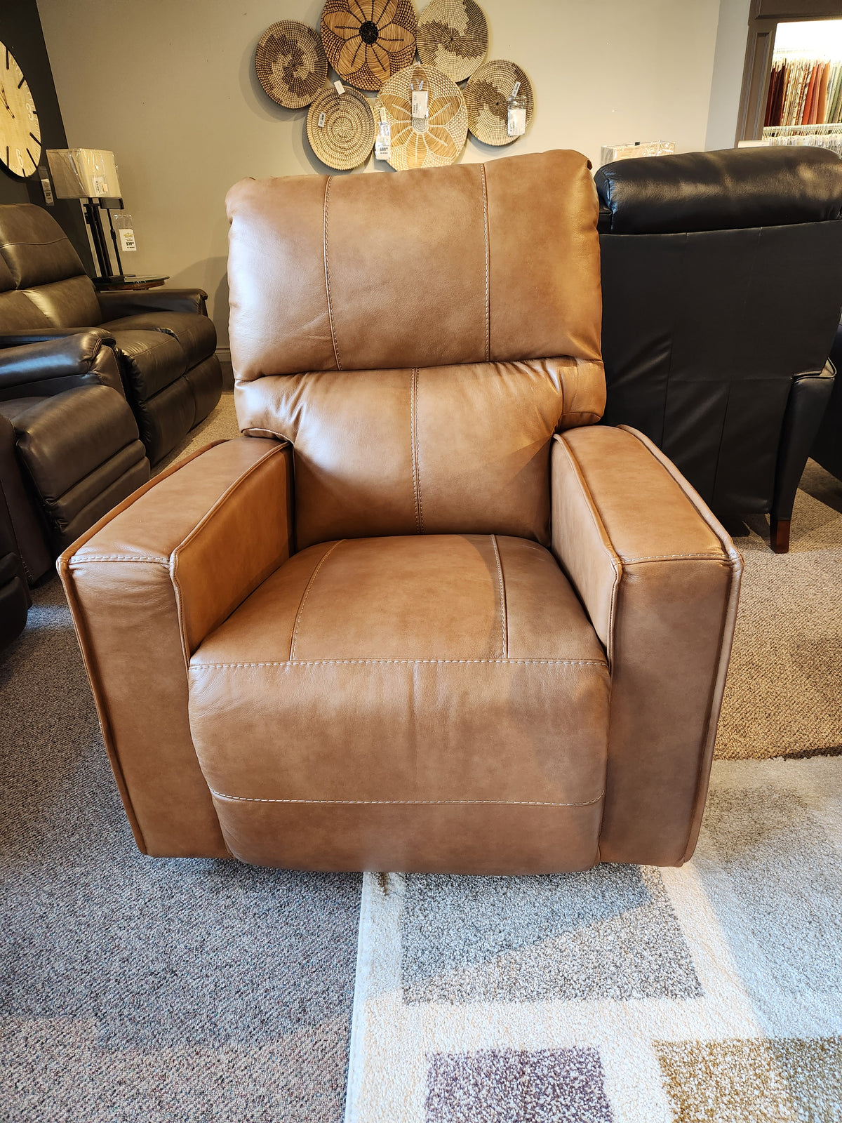 The La-z-boy 759 Maddox Rocker Recliner, offering style and comfort, is showcased in a furniture showroom with other seating in the background. Its modern touch enhances the charm of this exquisite brown leather piece.