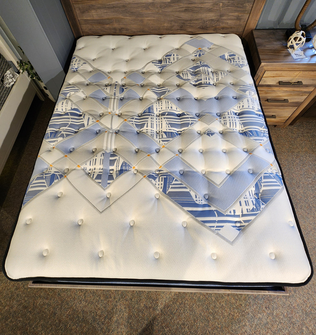 A neatly made bed with a blue and white geometric-patterned Jacob ET Soft Mattress from Sealy in a wooden frame, enhanced by the exceptional support of Sealy Comfort gel, sits elegantly next to a wooden nightstand.