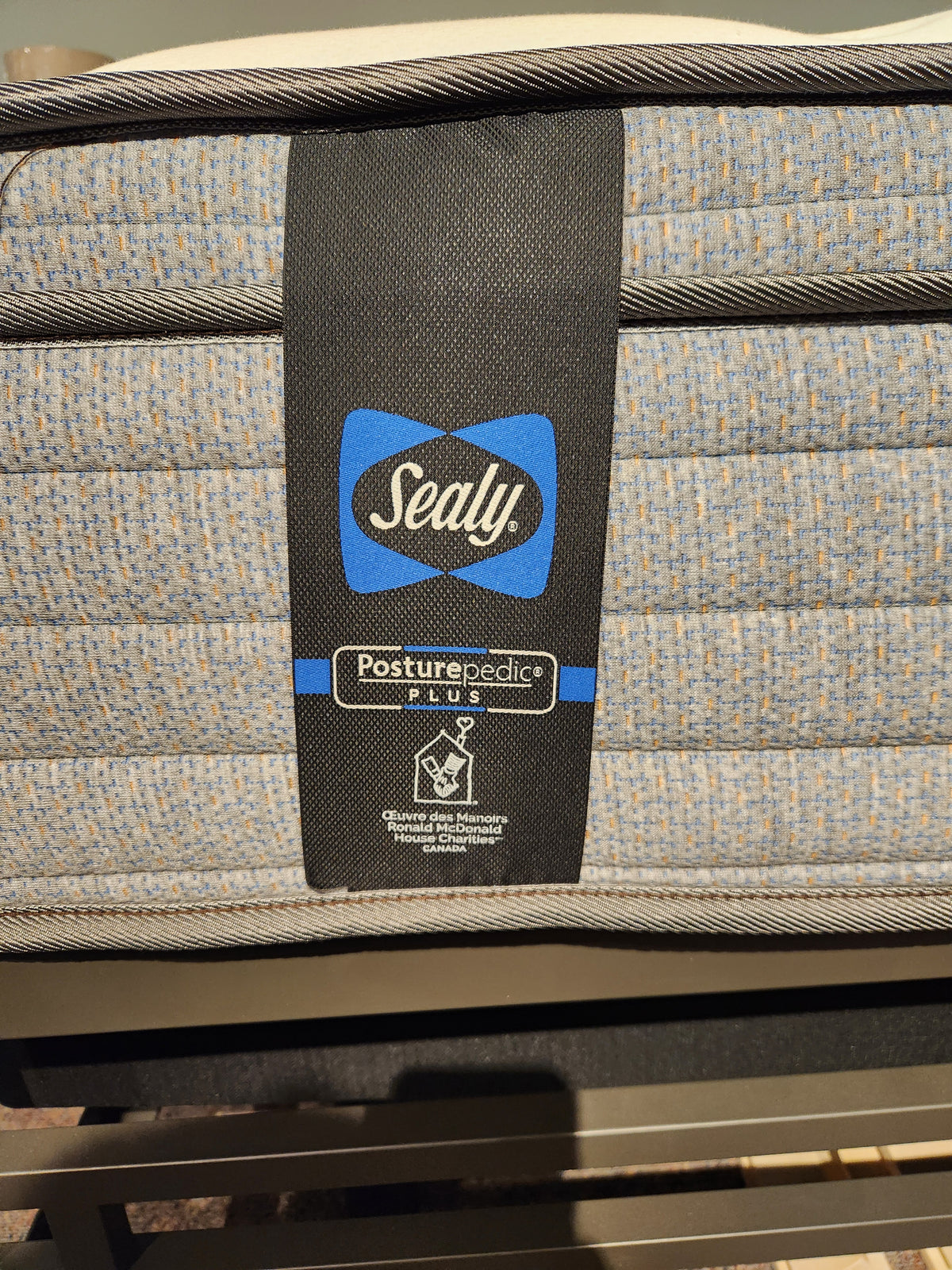 Close-up of a Sealy Callum ET Medium Mattress label, showing blue and gray textured fabric with Sealy Comfort gel.