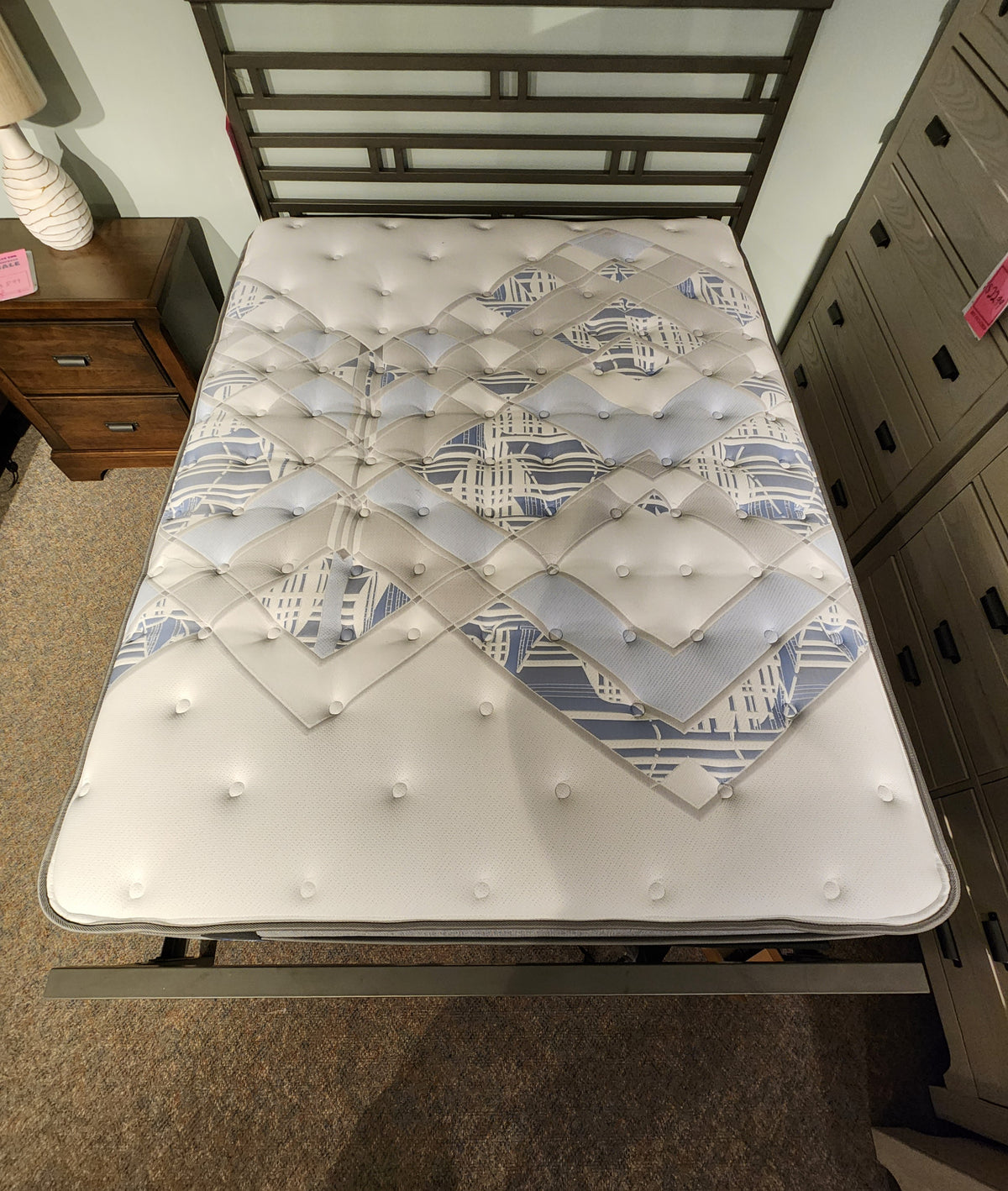 A Sealy Callum ET Medium Mattress with a geometric design on a metal bed frame, placed between nightstand and dresser.