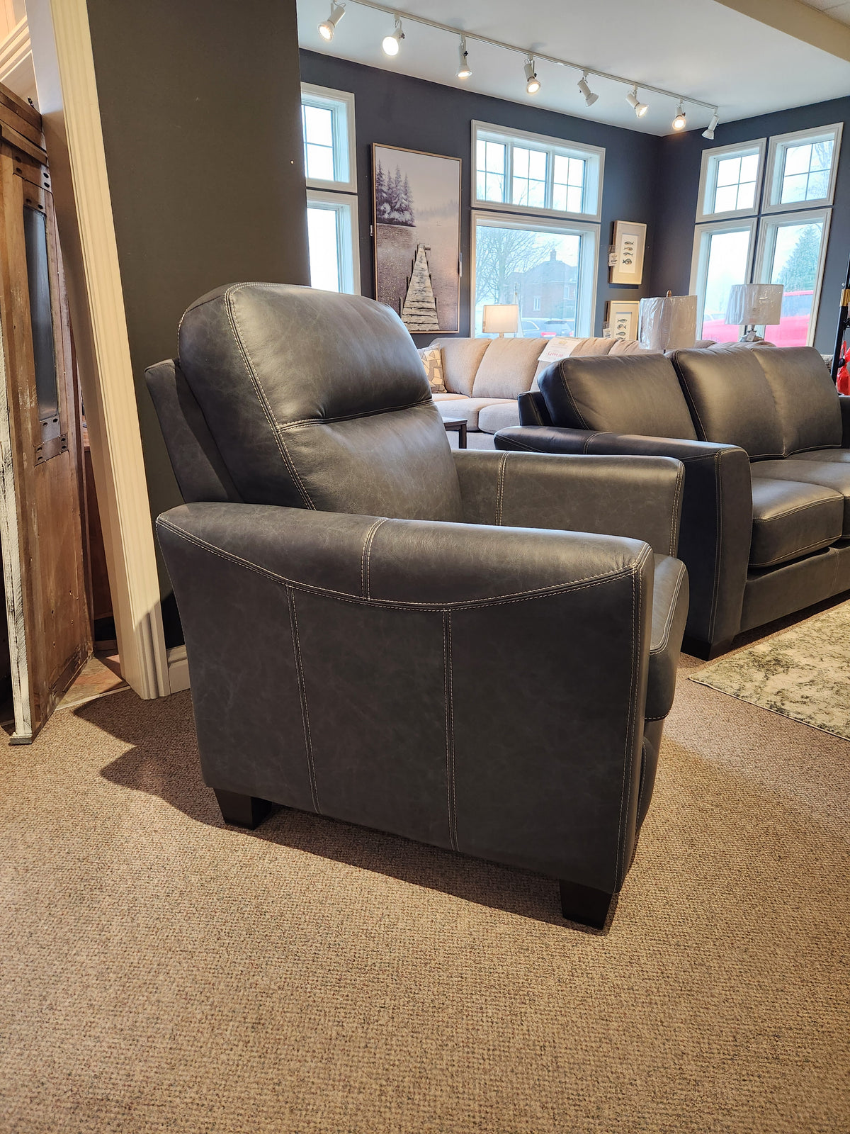 A 622 Leather Reclining Chair by Leather Craft in dark gray genuine leather is positioned in a showroom, with a sofa and large windows in the background.