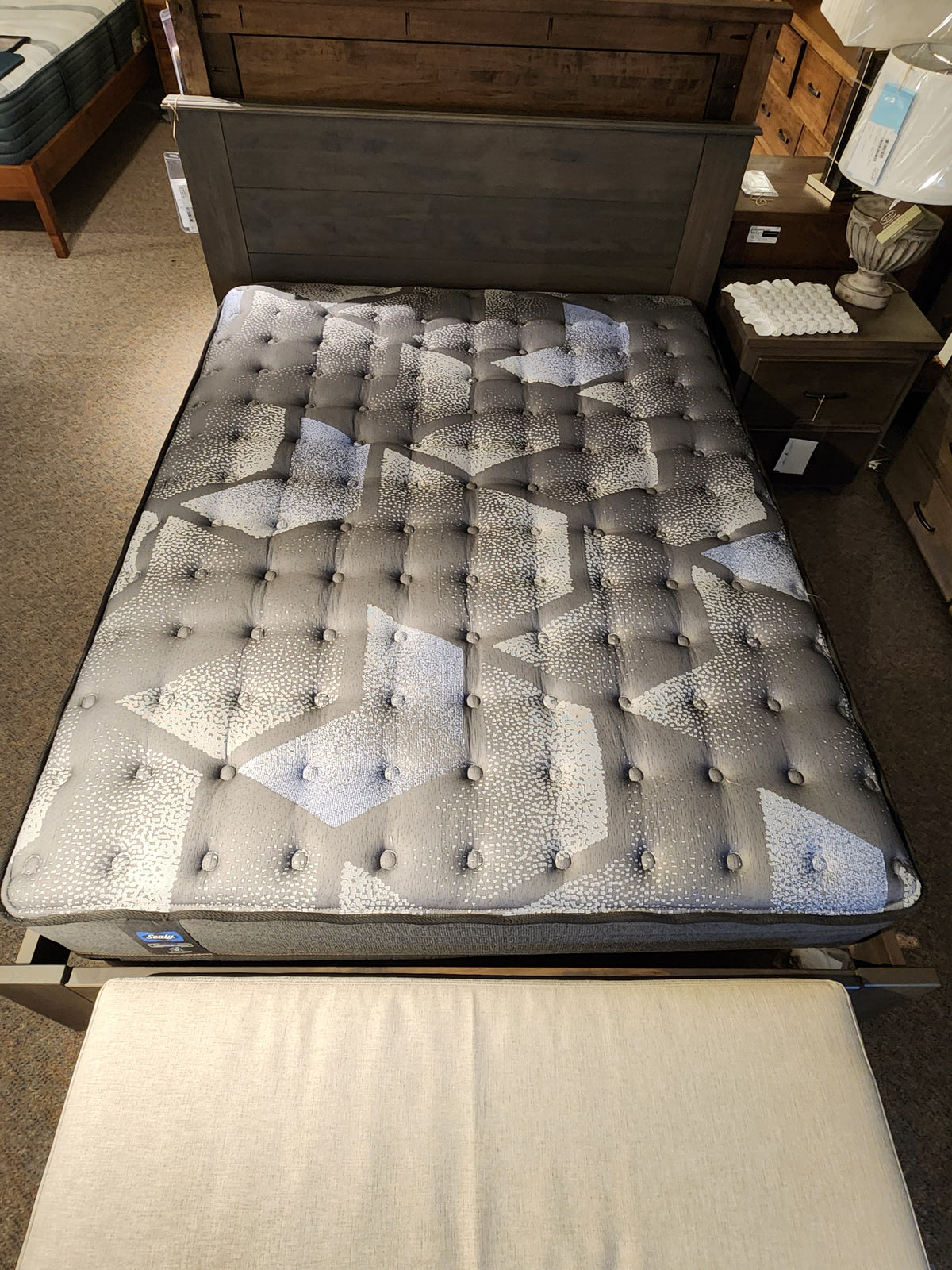 The Jaxon TT Firm Mattress by Sealy, displayed on a bed frame in the showroom, features a tufted design with geometric patterns. The headboard is made from wood and includes Certipur-US certified Sealy Comfort gel HD foam for exceptional support and luxury.