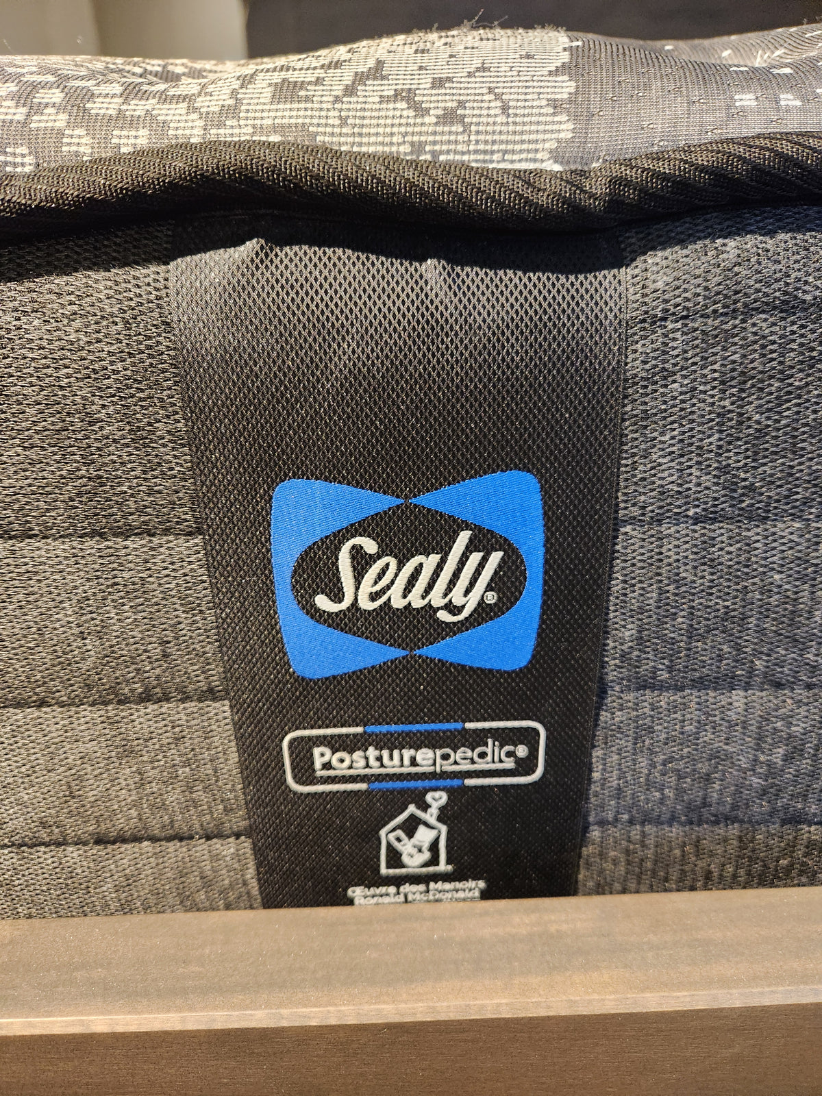 A detailed view of the label on the Sealy Jaxon TT Firm Mattress highlights its blue and white design set against a textured gray fabric. This mattress features Sealy's Comfort gel HD foam and holds a Certipur-US certification, offering both restful and eco-friendly sleep.