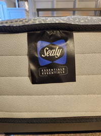 A close-up of the Sealy Nessa TT Medium Mattress label displays the word "Essentials." The mattress, Certipur-US certified, features gray fabric with a black border and is crafted for optimal pressure relief.