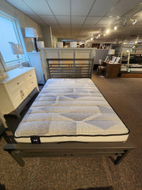 A showroom showcases a Sealy Nessa TT Medium Mattress with a striped design, resting on a metal frame and complemented by a wooden headboard. This Certipur-US certified mattress ensures outstanding pressure relief, enhanced by accompanying dressers and lamps for an inviting presentation.