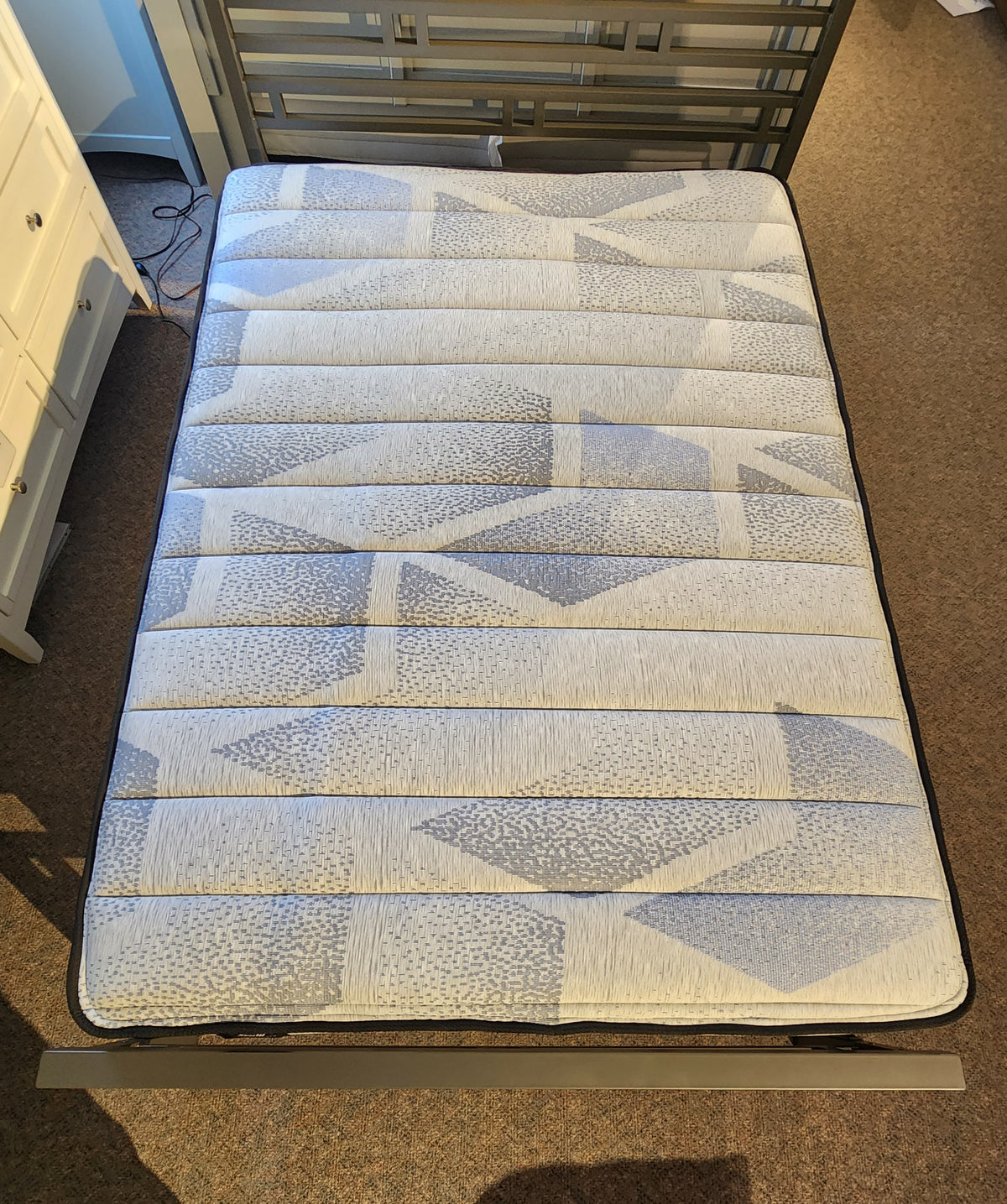 A twin-sized Nessa TT Medium Mattress by Sealy, celebrated for its pressure relief and Certipur-US certification, flaunts a geometric blue and white pattern on a gray bed frame in the carpeted room.