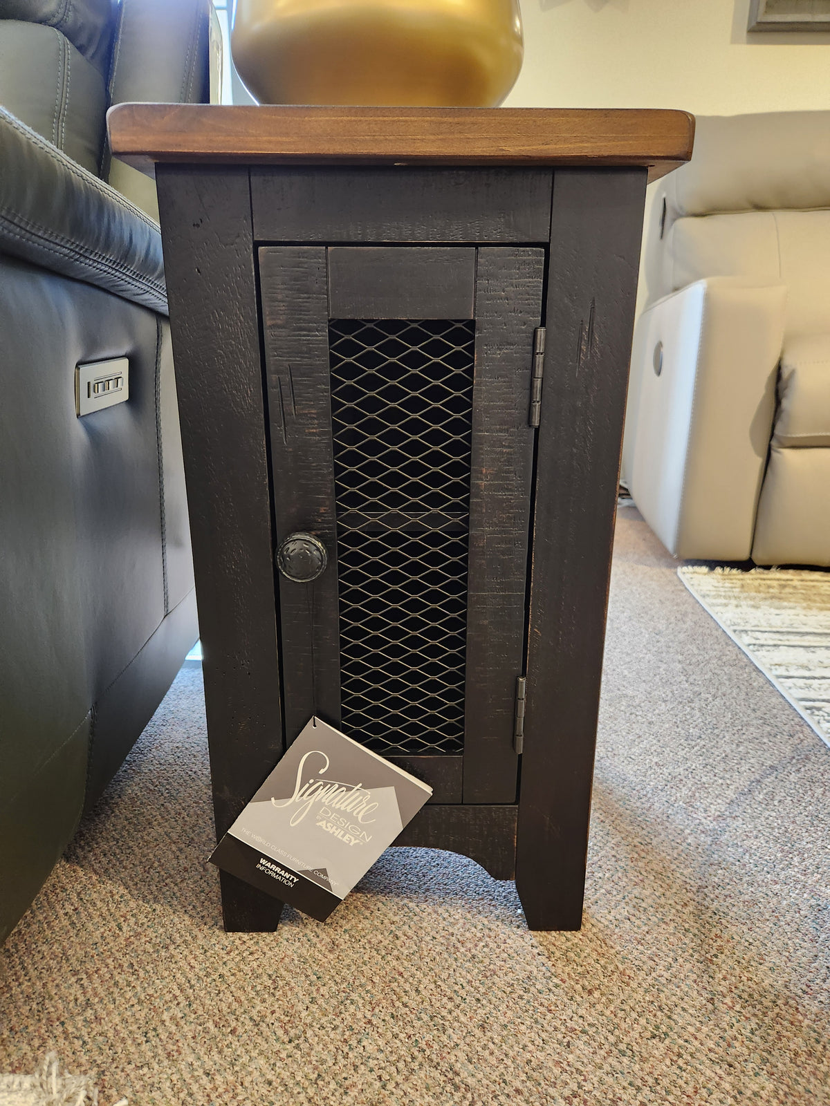 The Valebeck Chairside T468 End Table by Ashley, a small black wooden table with a mesh front, sits between couches.