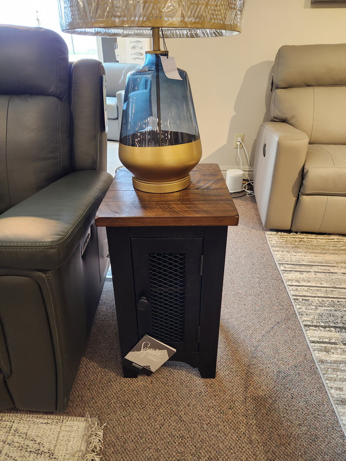 The Valebeck Chairside T468 End Table by Ashley with a blue and gold lamp stands between two recliners on carpet.