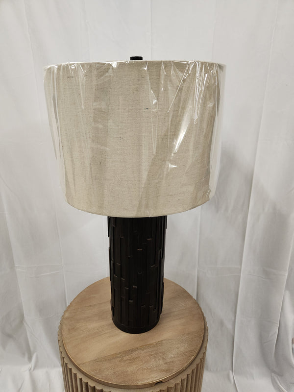 The Ashley L243234 Makya Table Lamp with a beige shade is wrapped in plastic on a wooden table for contemporary homes.