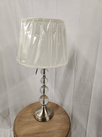 The Venzia Lamp by Renwil, showcasing a satin nickel finish, silver base, and white shade, is wrapped in protective plastic. It sits elegantly on a round wooden table against a pristine white curtain.