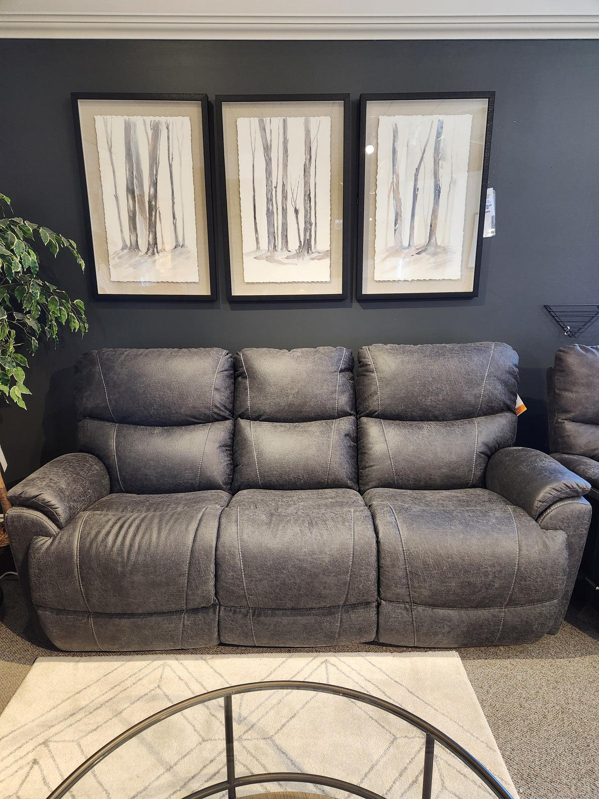 A gray 724 Trouper Power Reclining Sofa with a headrest by La-Z-Boy, featuring USB ports, stands against a dark wall, complemented by three framed tree artworks above. In front, a round glass coffee table completes the stylish setup.