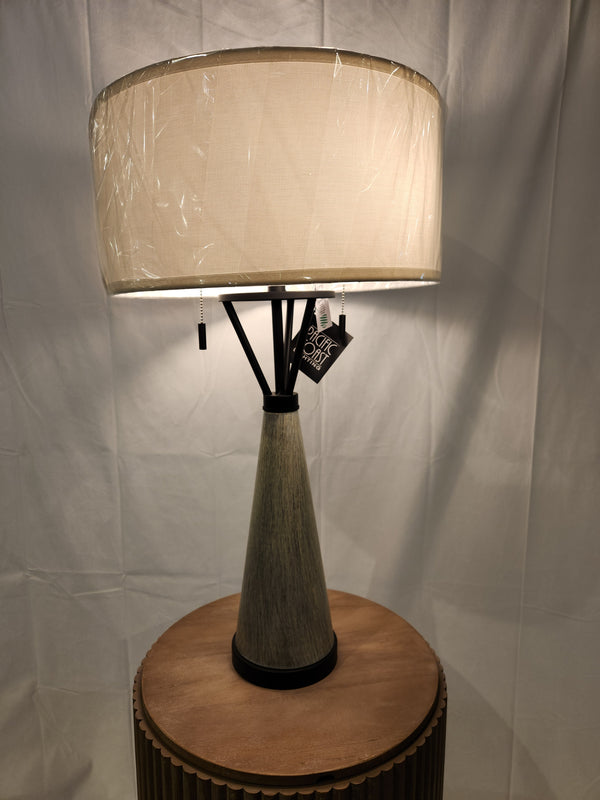 The 71N94 Oakland Table Lamp by Pacific Coast radiates industrial charm, featuring a cone-shaped base and a spacious beige fabric lampshade as it elegantly rests on a round wooden surface.