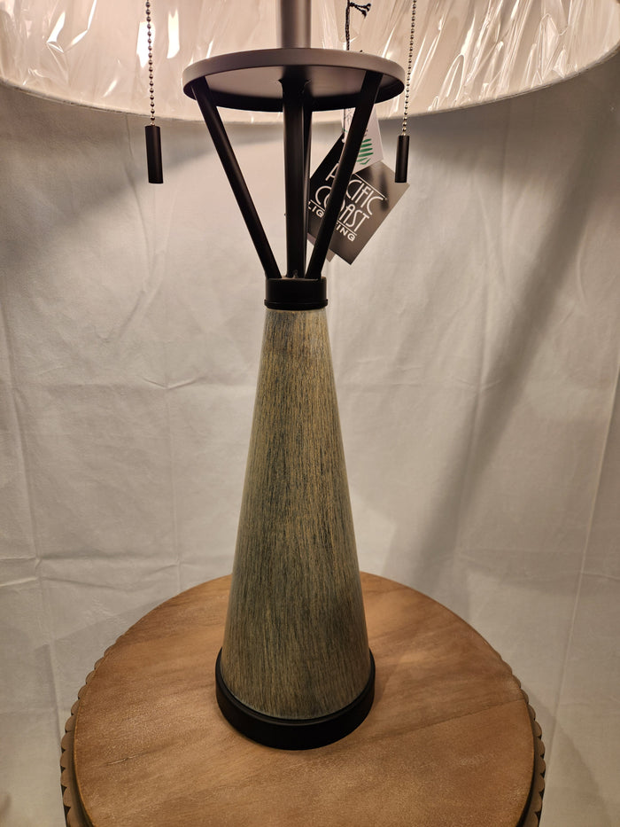 The 71N94 Oakland Table Lamp from Pacific Coast showcases a stylish design with its wooden conical base and triangular black metal frame, exuding industrial charm. It rests on a wooden surface beneath a white lampshade.