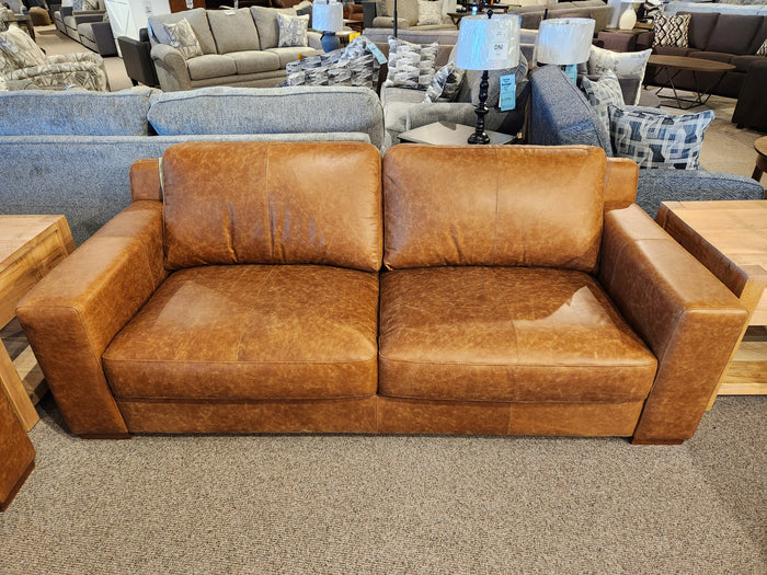 A Bowen Leather Sofa from Primo, crafted from top grain leather in a luxurious whiskey color, is spotlighted in a furniture showroom. It's nestled among other couches and complemented by a wooden side table, captivating attention with its premium finish and timeless style.