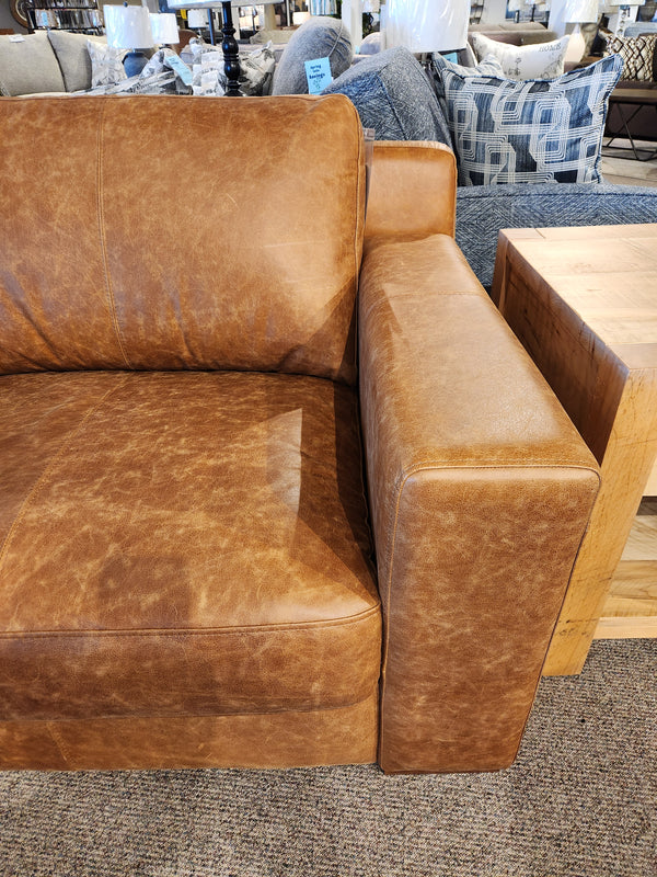 The Primo Bowen Leather Sofa in a luxurious whiskey hue boasts visible stitching detail. It is beautifully positioned beside a wooden side table on the carpeted floor, highlighting its top grain leather craftsmanship.