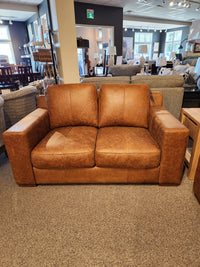 The Bowen Leather Loveseat by Primo, with its luxurious whiskey-colored top grain leather, is elegantly displayed in a furniture showroom alongside gray couches and wooden tables.