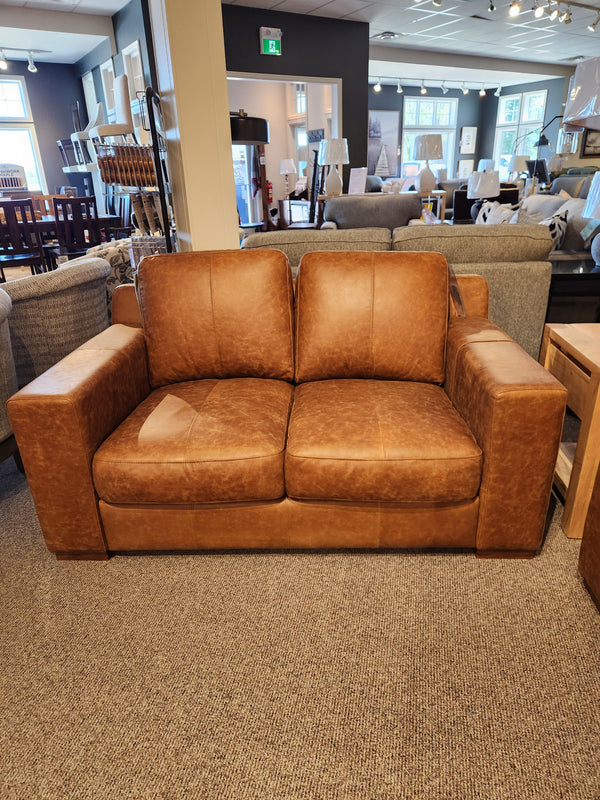 The Bowen Leather Loveseat by Primo, with its luxurious whiskey-colored top grain leather, is elegantly displayed in a furniture showroom alongside gray couches and wooden tables.