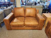 The Primo Bowen Leather Loveseat, made from premium top grain leather in a luxurious whiskey hue, is elegantly displayed among various furniture items on a soft carpeted floor.