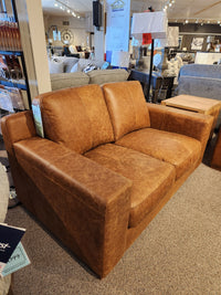 A Primo Bowen Leather Loveseat in a rich whiskey color is showcased in a furniture store, surrounded by other sofas and wooden side tables, highlighting its top grain leather craftsmanship.