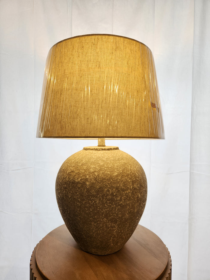 The Dreward Table Lamp by Ashley, with a beige shade and distressed gray finish, sits on a round wooden table.