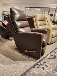 The 786 Liam Rocker Recliner by La-z-boy, crafted from rich brown leather and featuring a wooden lever, exudes casual style while sitting comfortably alongside other chairs in the furniture store showroom.