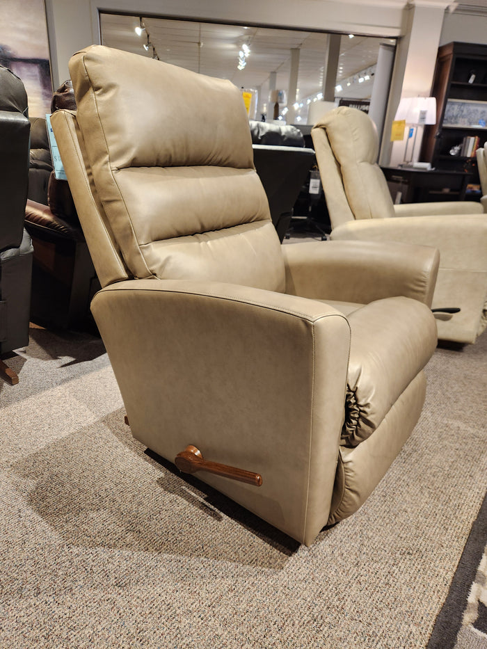 The 786 Liam Rocker Recliner by La-z-boy, boasting a casual design, is showcased in a furniture showroom. This beige recliner chair features a wooden handle on one side and offers exceptional comfort for any home setting.