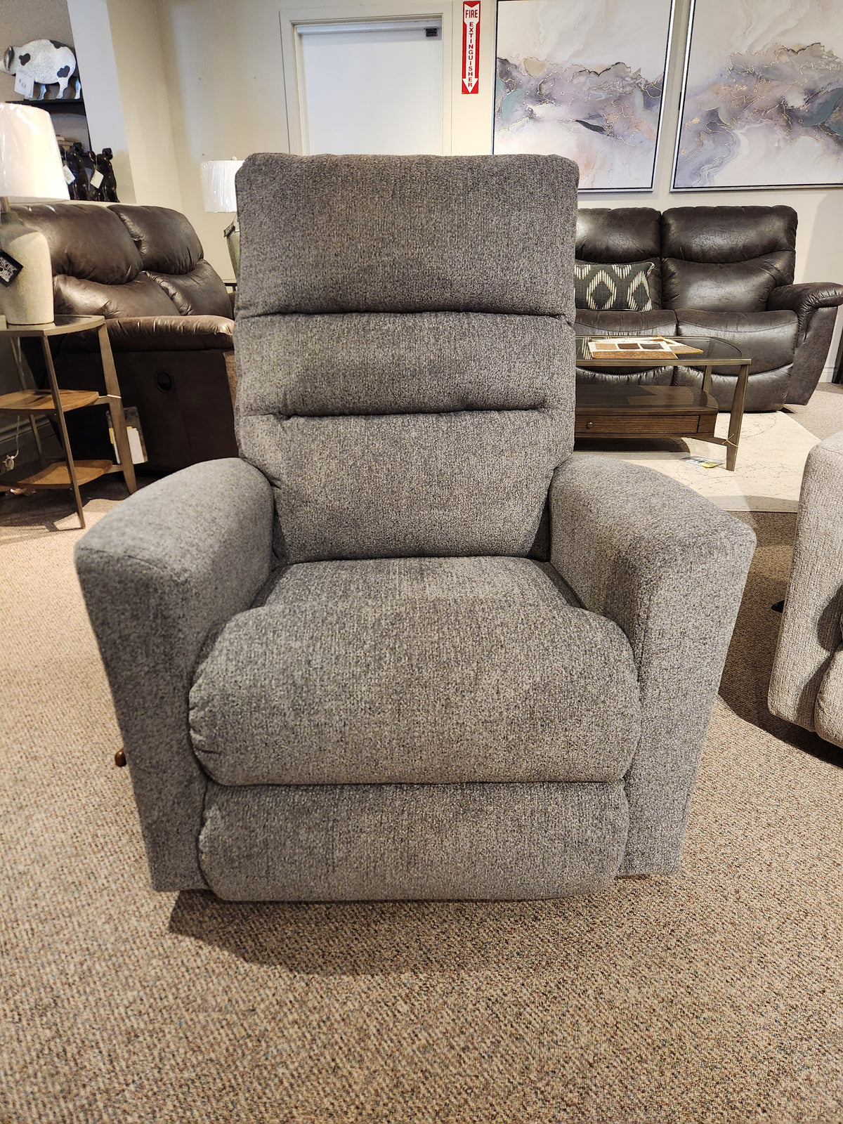 The 786 Liam Rocker Recliner by La-z-boy, featuring gray upholstery, is displayed in a showroom setting on a carpeted floor. Its casual style effortlessly merges comfort with elegance, making it an ideal addition to any living space.