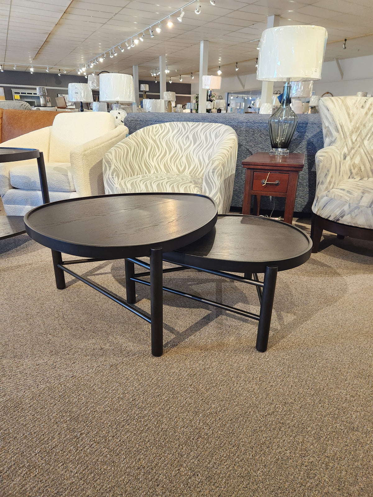 Displayed in a furniture showroom, the T5711-52 Hadleigh Coffee Nesting Table from Magnussen features a pair of round black tables with slender legs and a sleek coffee bean finish, perfectly embodying modern organic style among the surrounding chairs and lamps.