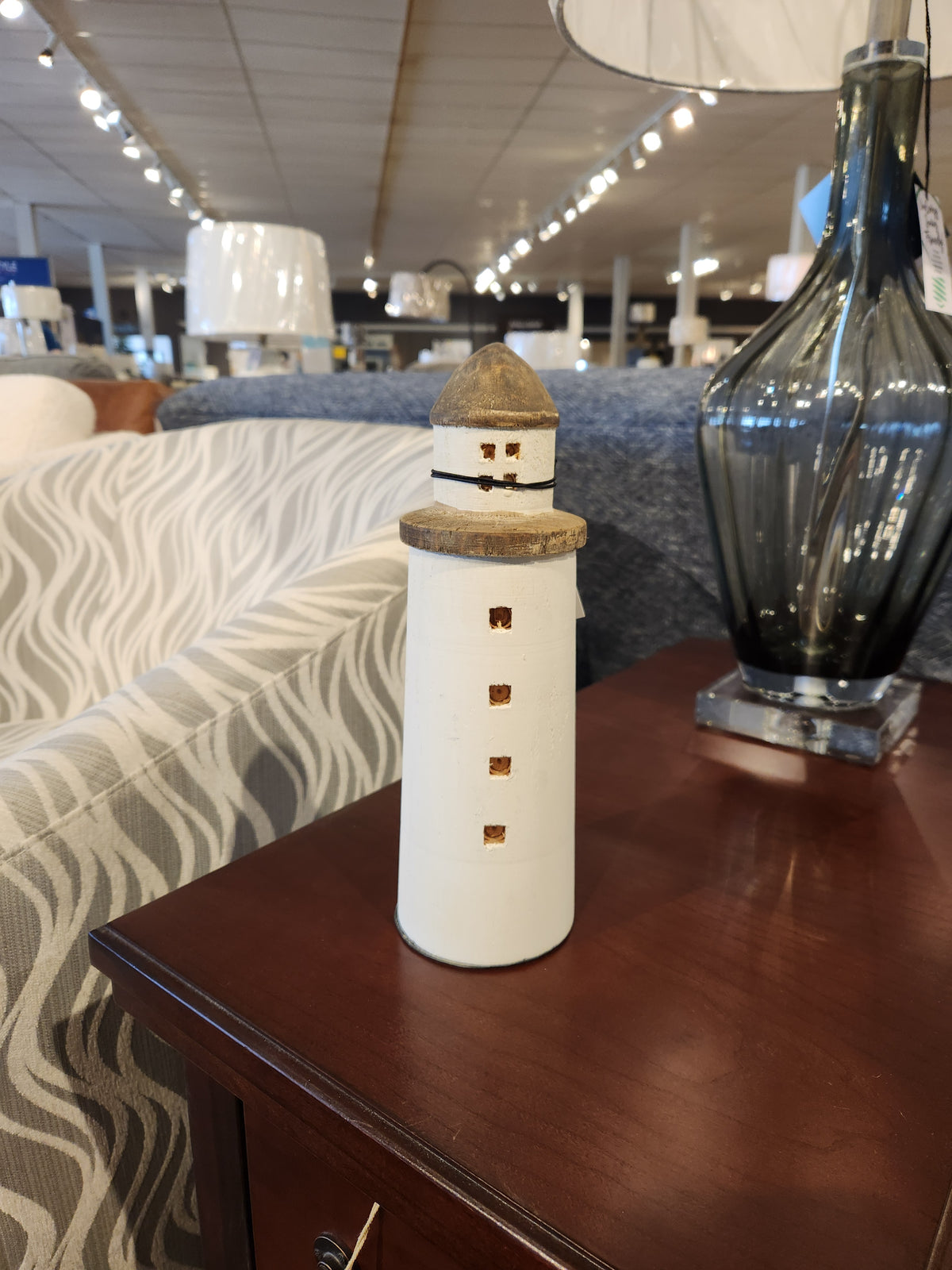 A coastal-inspired 68795 Lighthouse (Small) ornament from Mercana sits gracefully on a wooden table in a furniture showroom, accompanied by a lamp and a patterned sofa. This charming decorative object adds nautical charm to the setting.