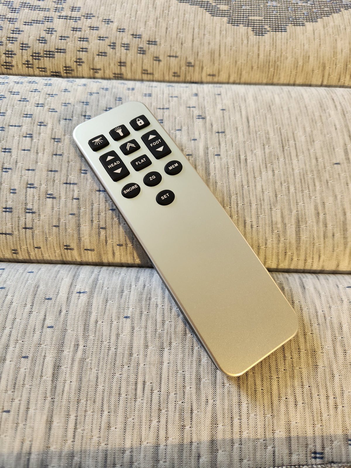 A remote control for the Essentials Adjustable Base by Fashion, featuring a sleek silver design with black buttons, is placed on a surface covered in light patterned fabric.