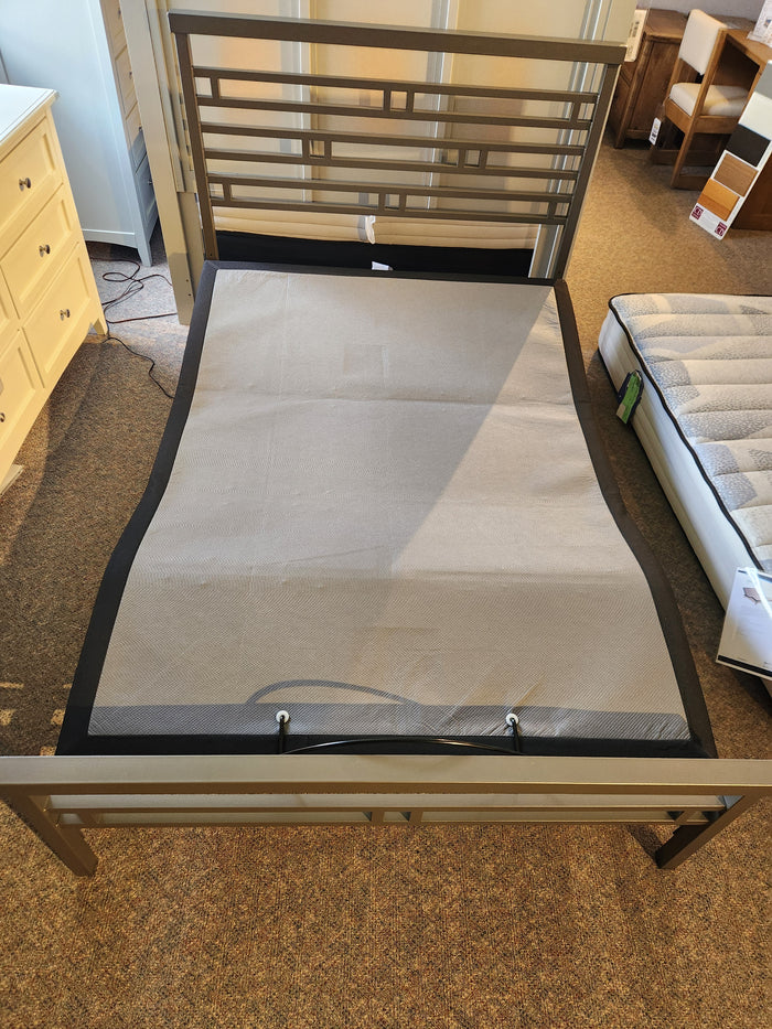 In a furniture showroom, an empty metal bed frame featuring a fabric support panel is showcased beside a mattress and dresser. The setup emphasizes the frame's compatibility with the Essentials Adjustable Base by Fashion, providing added convenience and modern functionality for your bedroom needs.