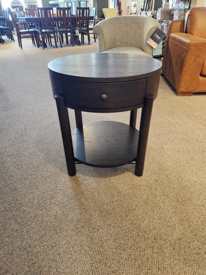 The T5711-05 Hadleigh Round End Table by Magnussen, with a drawer and shelf, shown in a carpeted showroom.