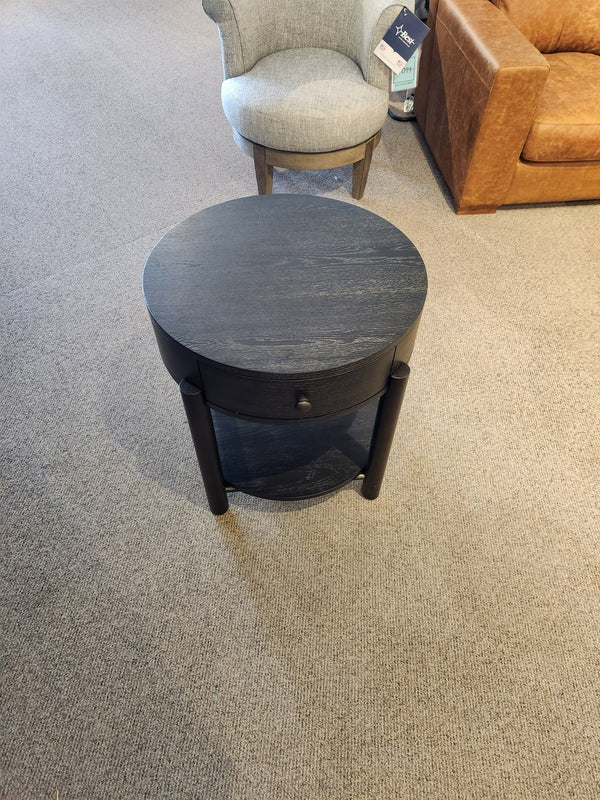 Magnussen's T5711-05 Hadleigh black round end table with drawer, near a blue chair on a carpeted floor.