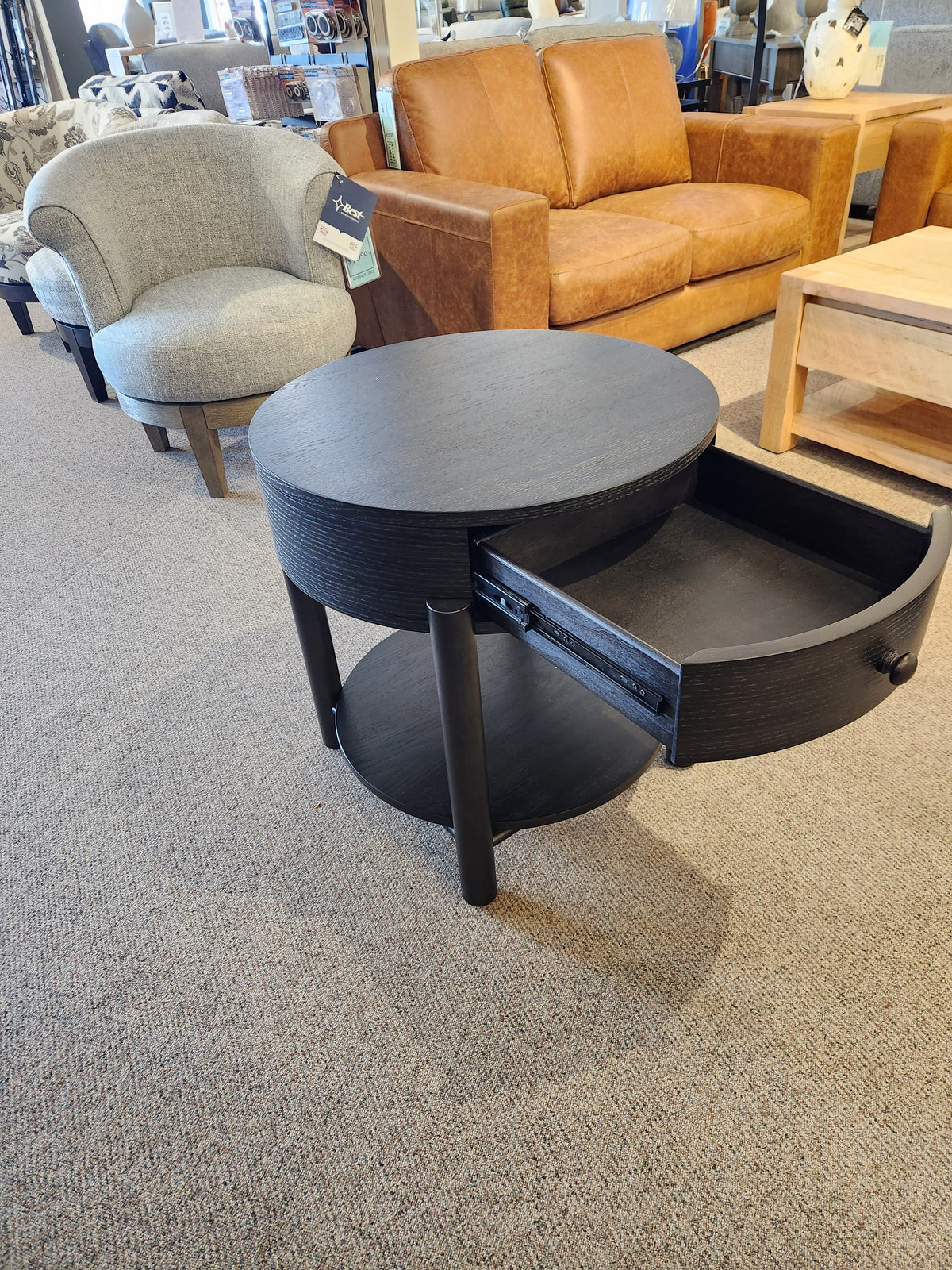 Modern organic showroom with a Magnussen T5711-05 Hadleigh Round End Table, black, open drawer, surrounded by sofas.