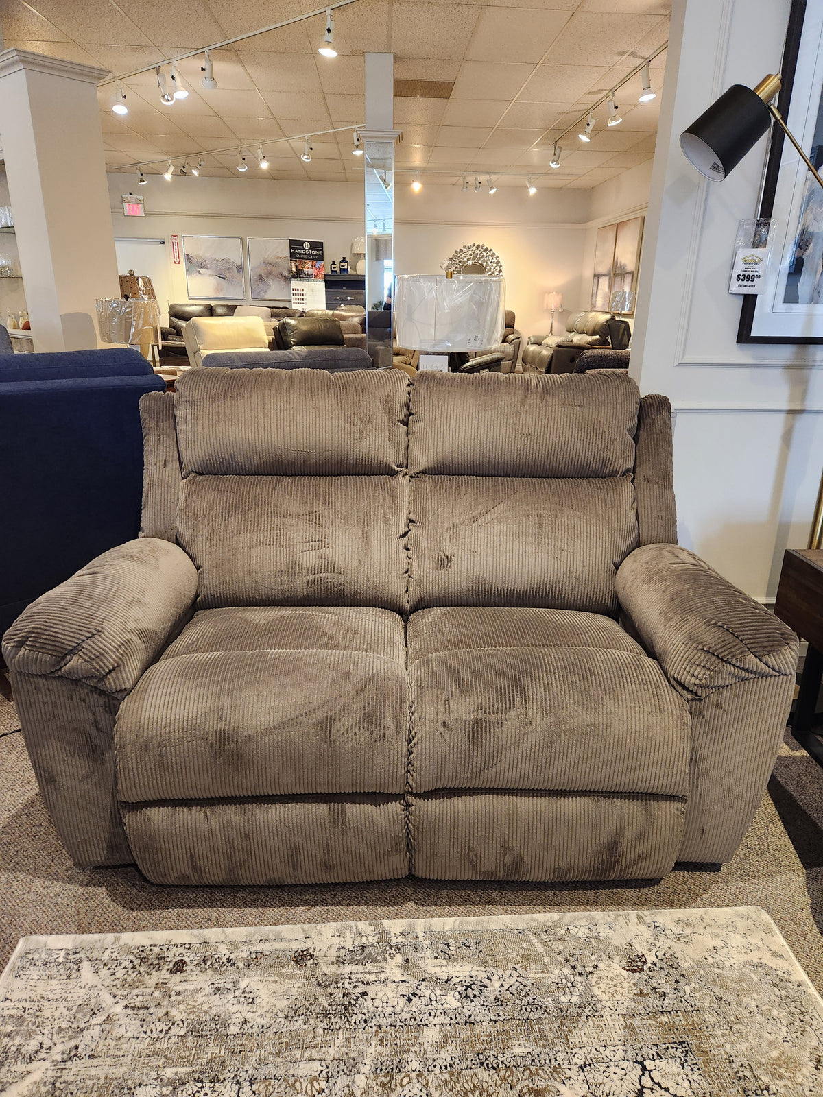 The La-z-boy 761 Joel Reclining Loveseat, featuring a plush brown corduroy texture and cushioned arm pads for inviting comfort, is showcased in a furniture showroom.