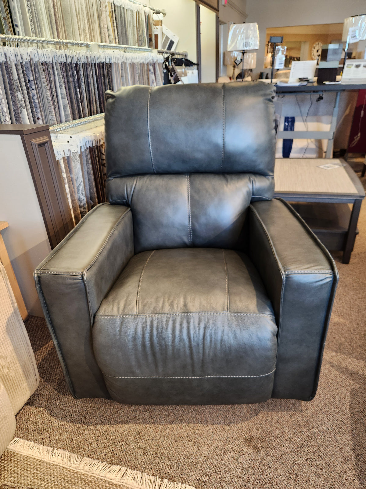 The 759 Maddox Rocker Recliner by La-z-boy, featuring a luxurious dark green leather finish, offers an exceptional combination of style and comfort. Presented in a furniture showroom with various fabric samples and a side table, it effortlessly enhances the modern appeal of any room.