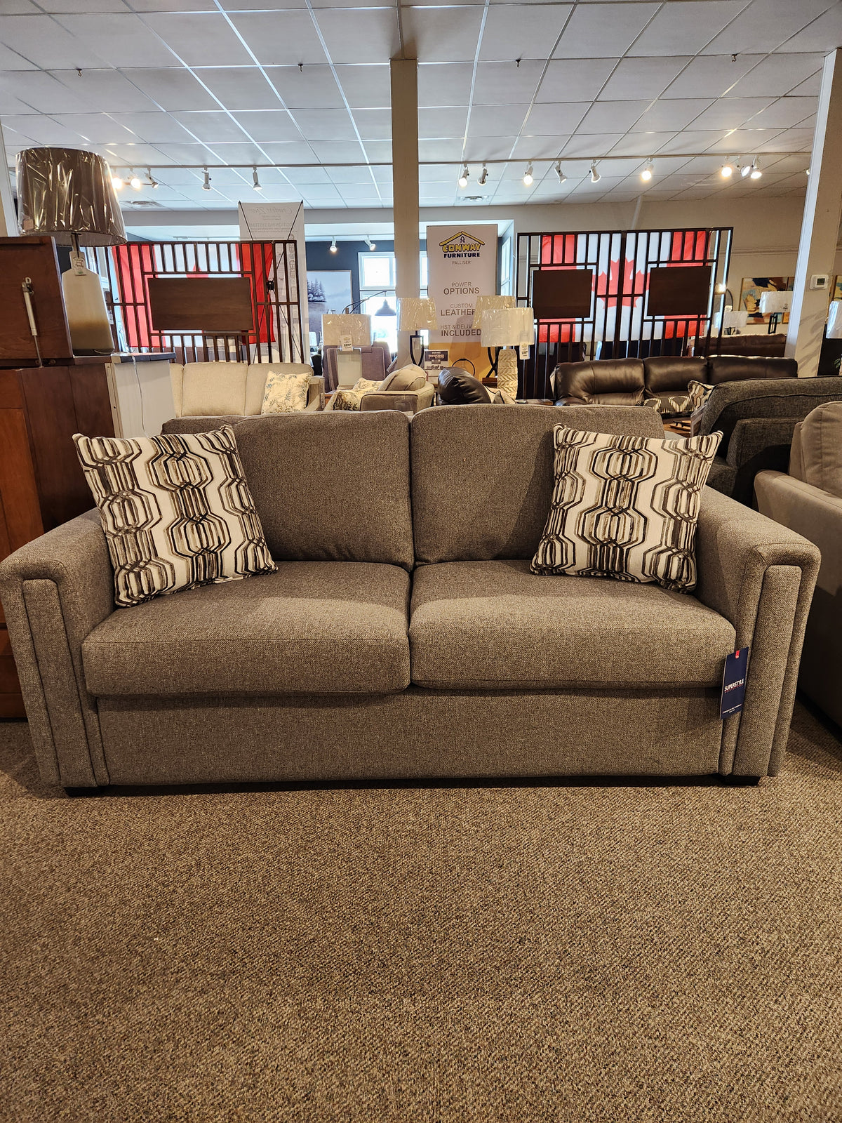 The Simmons Upholstery showroom is featuring the 9350 Double Sofa Bed, a gray fabric piece with sleek track arms and two patterned pillows.