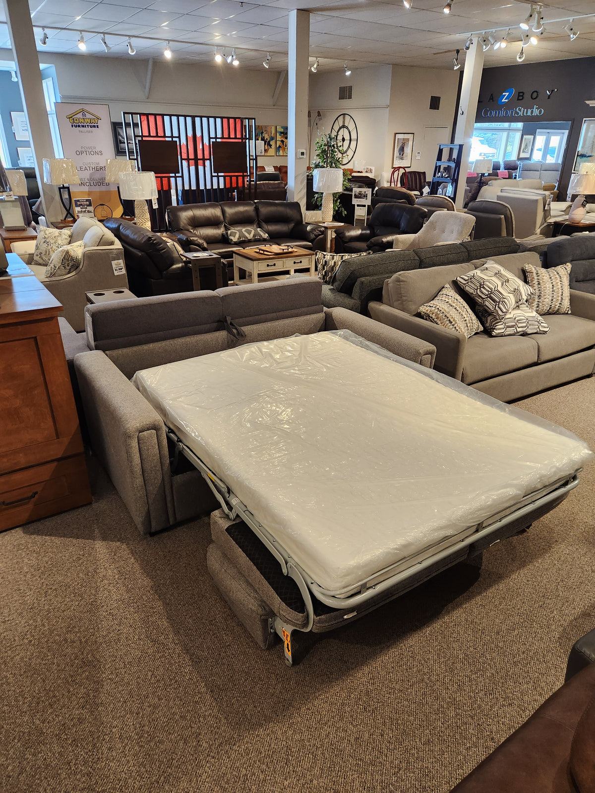 A 9350 Double Sofa Bed from Simmons Upholstery, featuring a sleek grey design with track arms and an extended mattress, is showcased in a furniture showroom. Numerous other sofa beds and decor items can be seen in the background, adding to the cozy atmosphere.