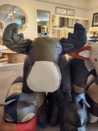 Plush toys shaped like a moose and various other animals are showcased alongside Reiner's Leather Animal Ottomans by Reiner's Originals in a store showroom, with fabric samples displayed in the background.