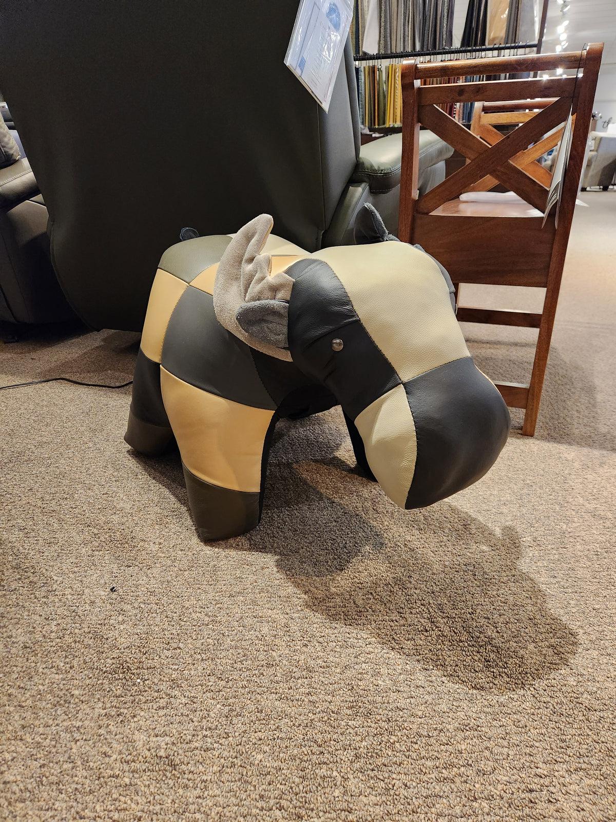 This distinctive piece from Reiner's Originals, the Reiner's Leather Animal Ottoman in a rhino shape, boasts a patchwork design and stands proudly on the carpeted floor beside a wooden chair.