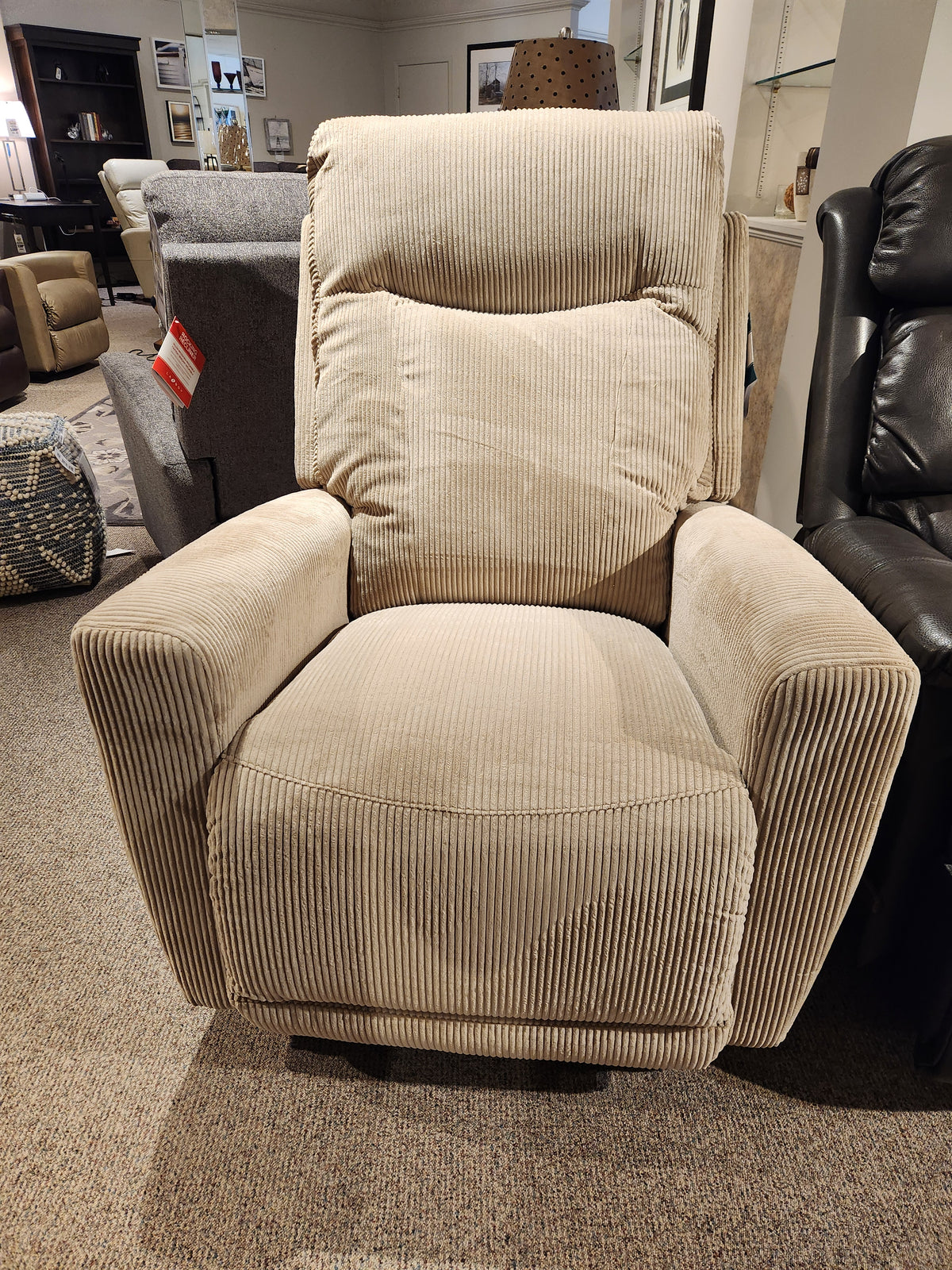 The 715 Kodie Rocker Recliner by La-z-boy, made from luxurious corduroy fabric, rests comfortably on the carpeted floor of the store, providing a perfect place for ultimate relaxation.