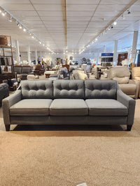 The 77190-01 Tristan Sofa by Palliser, a stationary three-seater in stylish gray, is displayed in a furniture showroom among a variety of diverse pieces. Its deep seating offers an invitation to relaxation and comfort.