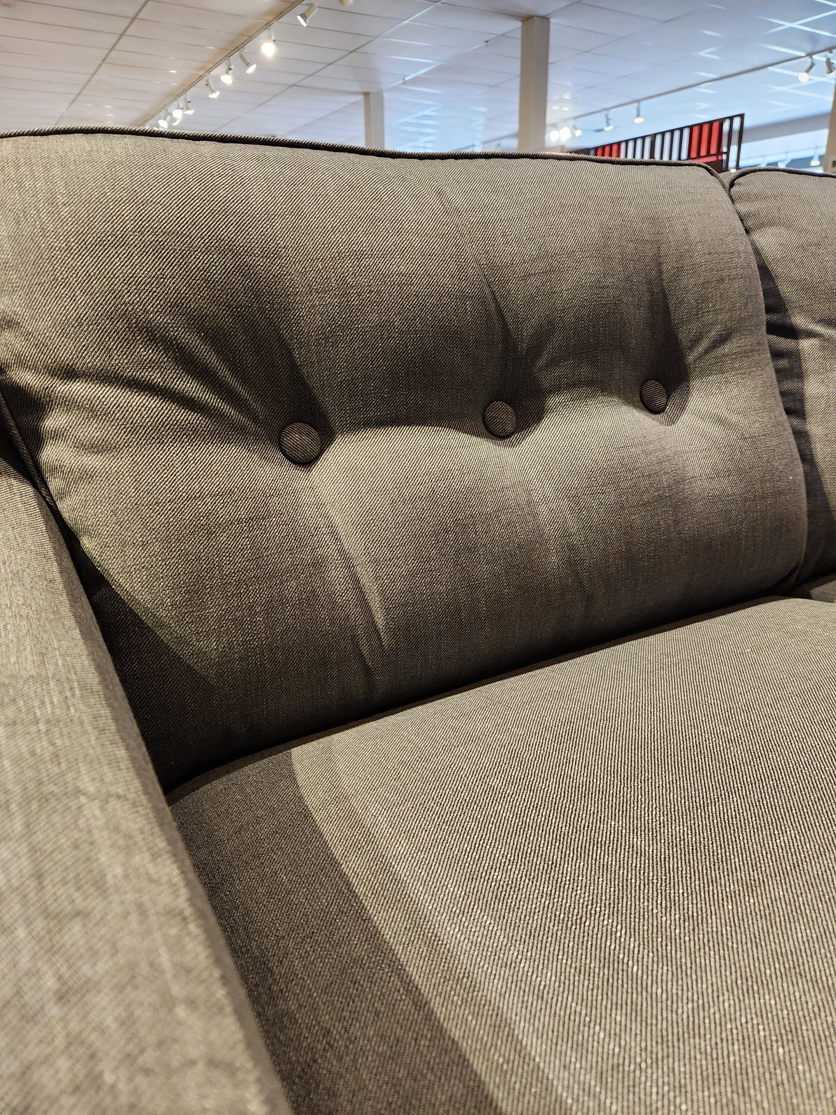 Close-up of the Palliser 77190-01 Tristan Sofa, showcasing its gray upholstered design with button-tufted back cushions and deep seating comfort under showroom lighting.