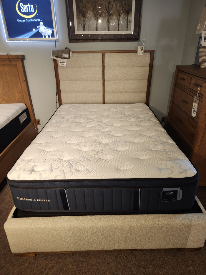 A neatly made bed showcasing a Stearns & Foster Amalia Pillow Top Cushion Firm Mattress with a beige headboard, beside a wooden nightstand in a showroom, highlighting exceptional breathability.