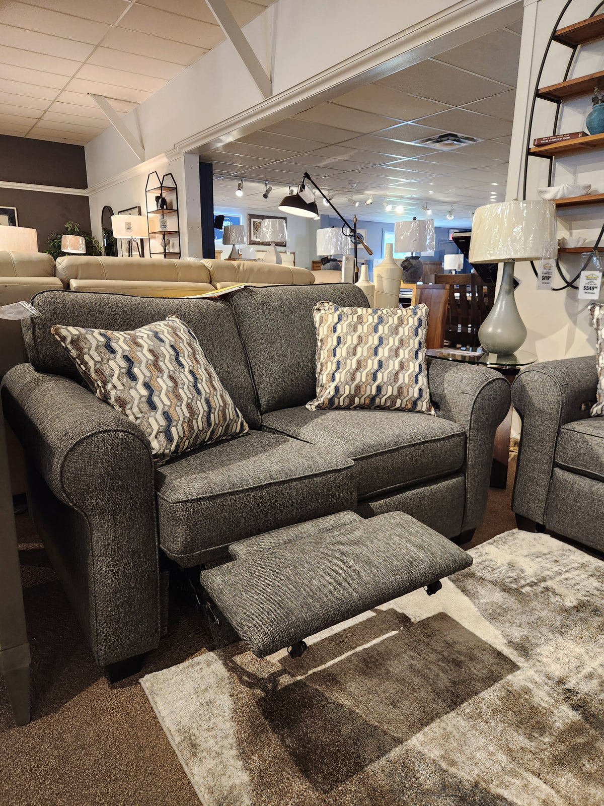 The 2179 Power Loveseat by Decor-rest, featuring gray fabric with geometric-patterned pillows and an extended footrest, showcases exquisite Canadian-made upholstery in a furniture showroom.