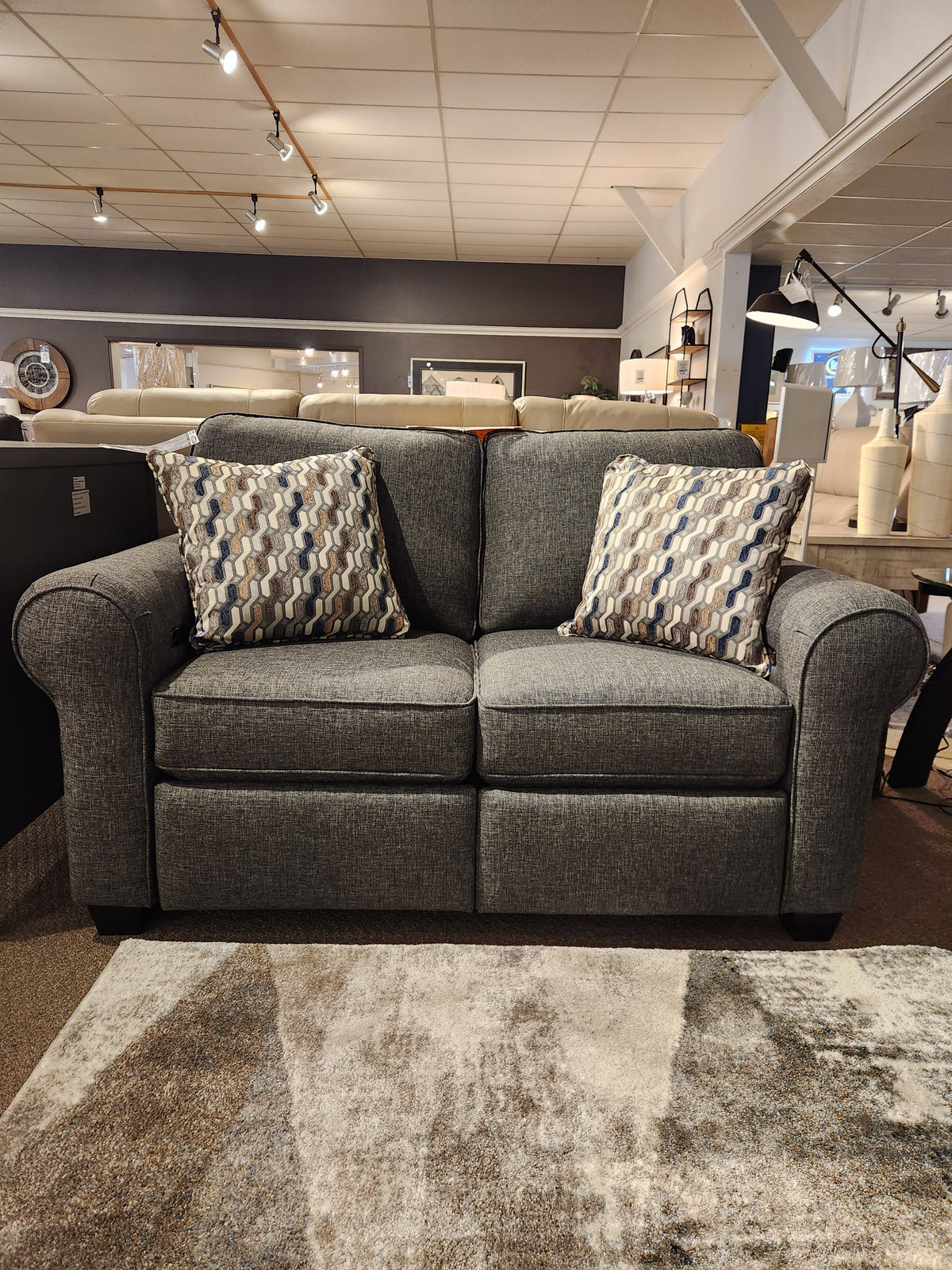 The Decor-rest 2179 Power Loveseat, a Canadian gray upholstered piece complete with two patterned pillows, is elegantly displayed in a showroom.